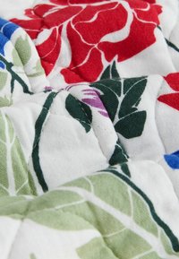 Steppstoff mit gesteppten Blumenmustern in Rot, Grün, Blau und Lila auf weißem Hintergrund, der Textur und Falten zeigt.