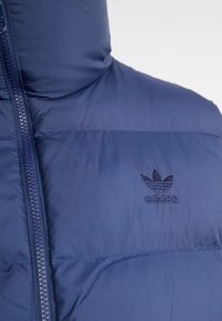Marinblå Adidas vadderad jacka med hög krage och synlig framsida dragkedja, med broderad Adidas-logotyp på bröstet.