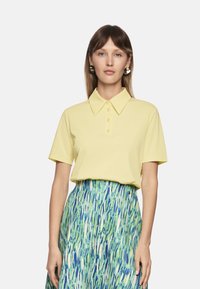 Gelbes Poloshirt mit Kragen und Knopfleiste, kombiniert mit einem gemusterten grünen und blauen Rock mit vertikalen Streifen.