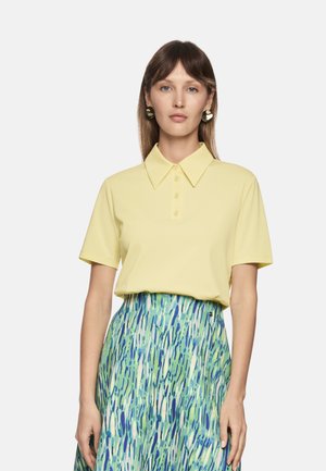 Gelbes Poloshirt mit Kragen und Knopfleiste, kombiniert mit einem gemusterten grünen und blauen Rock mit vertikalen Streifen.