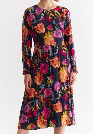 Frau trägt ein knielanges, langärmeliges Kleid mit dunklem Hintergrund und einem leuchtend orange-, pink- und violetten Blumenmuster.