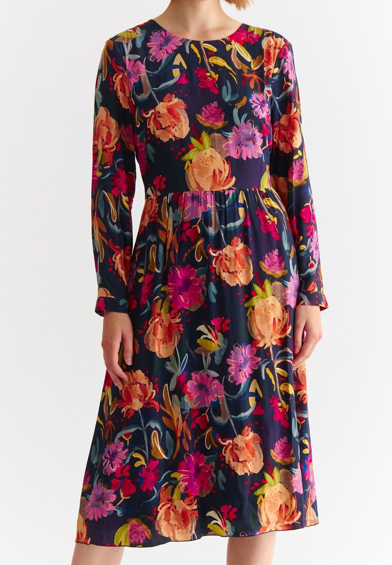 Femme portant une robe longue aux manches longues, longueur genou, avec un fond sombre et un motif floral aux couleurs orange vif, rose et violet.