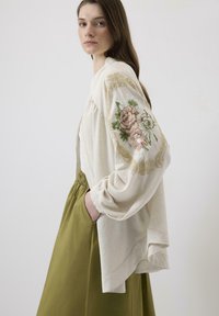 Cardigan supradimensionat de culoare crem, cu broderie florală în nuanțe de roz și verde pe un mânecă, asortat cu o fustă verde măsliniu, cu talie înaltă.