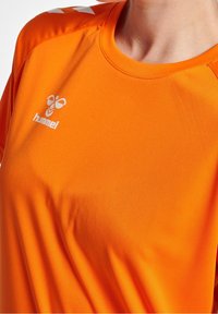 Jasnopomarańczowa koszulka sportowa wykonana z gładkiego materiału. Posiada okrągły dekolt oraz wyraźne białe logo i akcenty na ramionach.