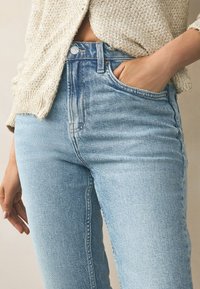 Světle modré džíny s vysokým pasem a přiléhavým střihem, vybavené zapínáním na knoflíky a bočními kapsami. Textura je mírně opotřebovaná, s viditelným šitím.