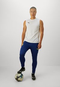 Άσπρο αμάνικο μπλουζάκι Puma, μπλε εφαρμοστά παντελόνια με νεόν λογότυπο, μαύρες ποδοσφαιρικές μπότες, στή standing σε μια μαύρη και λευκή ποδοσφαιρική μπάλα.