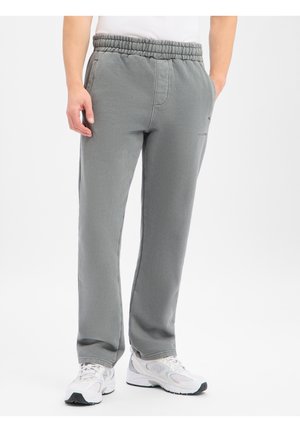 Man die grijze joggingbroek met elastische taille en witte sportschoenen draagt, staat met handen in de zakken tegen een witte achtergrond.
