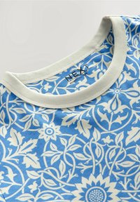 Niebieska koszulka z kwiatowym wzorem, z okrągłym, kremowym kołnierzykiem, wykonana z miękkiego materiału, z białymi konturami kwiatów i liści rozproszonymi po całej powierzchni.
