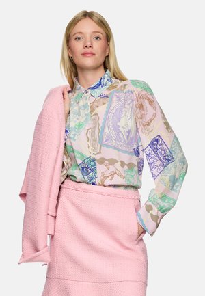 Blonde Frau trägt eine pastellfarbene, mehrfarbig gemusterte Bluse und einen rosa strukturierten Rock, hält eine passende rosa Jacke über der Schulter.