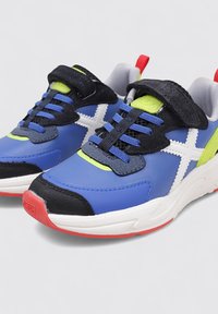 Zapatillas deportivas azules con acentos negros, logo blanco y detalles en verde brillante. Presentan materiales texturizados y una suela roja para un contraste adicional.