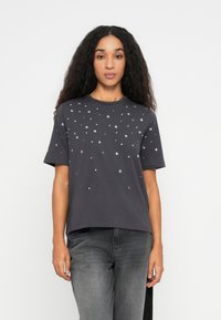 Ung kvinde med langt krøllet hår iført en mørkegrå T-shirt med spredte hvide stjerner og prikker, kombineret med falmede grå jeans.