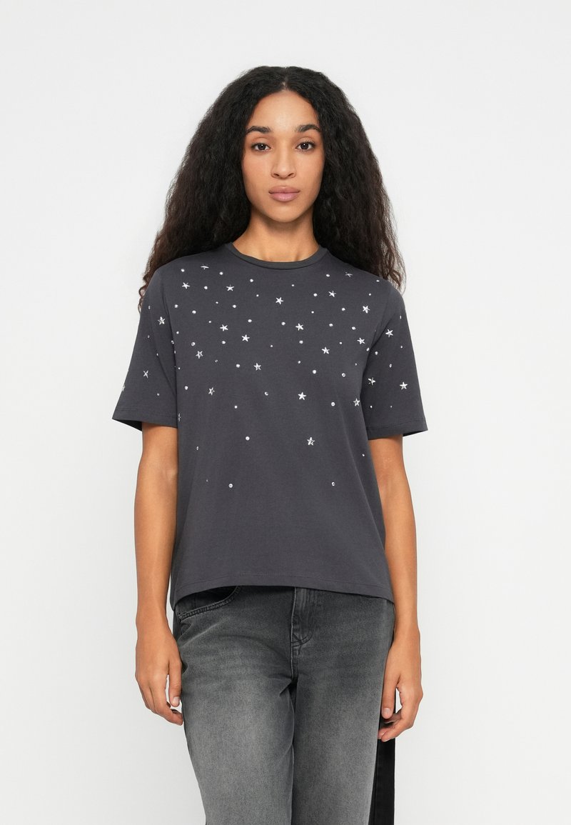 Ung kvinde med langt krøllet hår iført en mørkegrå T-shirt med spredte hvide stjerner og prikker, kombineret med falmede grå jeans.