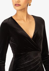 Schwarzes Samtkleid mit tiefem V-Ausschnitt, langen Ärmeln und Raffung an der Taille. Goldene Creolen ergänzen das Outfit.
