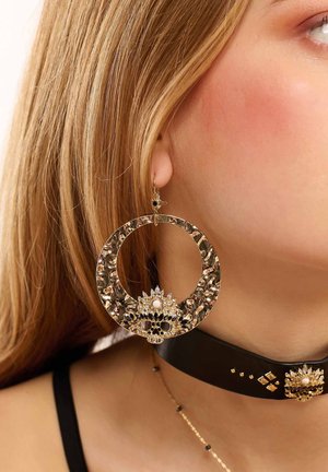 Goldene Creolen mit texturiertem Oberflächen, die am Boden ein mit Edelsteinen akzentuiertes schwarzes Totenkopfdesign und einen hängenden Verschluss aufweisen.