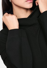 Schwarzer Pullover mit hohem Kragen, gerippten Details und weichem Material, mit nahtlosen Bündchen und einem dreieckigen Akzent am Kragen.