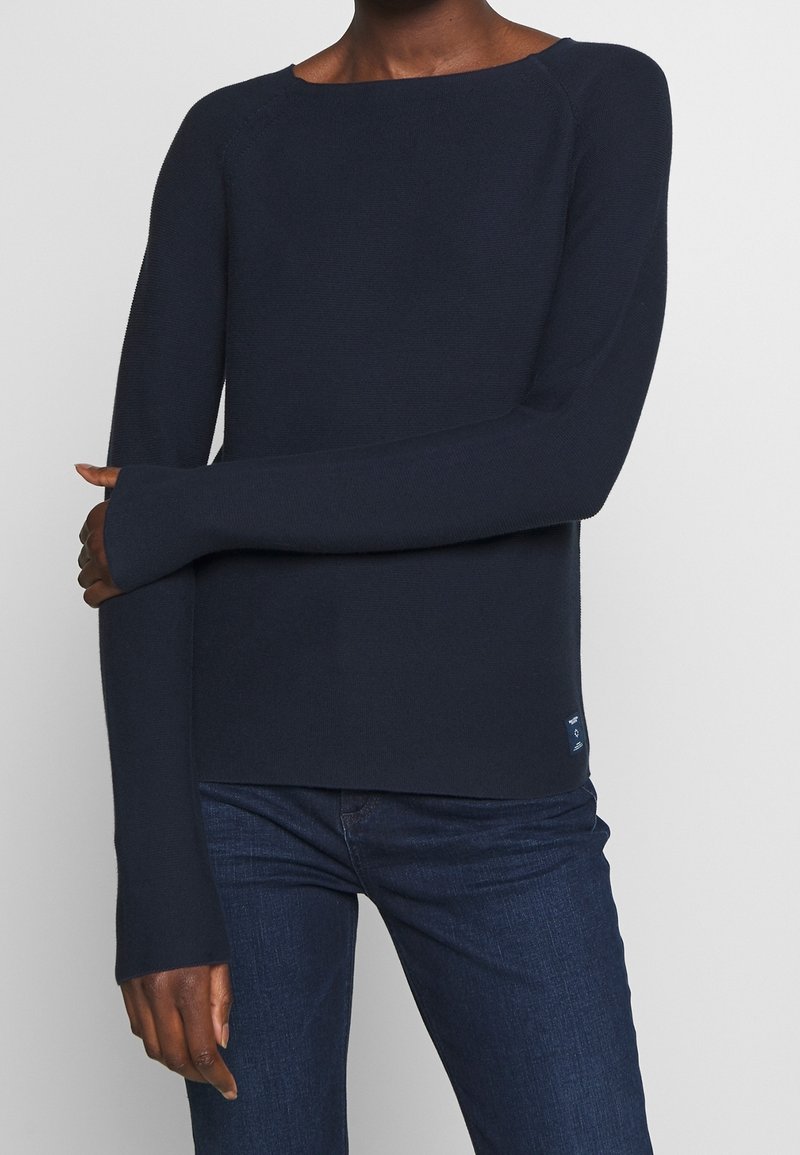 Personne portant un pull bleu marine à manches longues avec des trous pour les pouces et un jean bleu foncé, se tenant devant un fond clair uni.