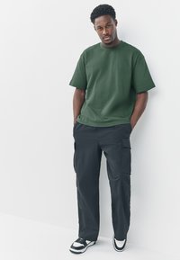 Grünes Baumwoll-T-Shirt mit rundem Ausschnitt, kombiniert mit schwarzen Cargo-Hosen mit Seitentaschen. Dazu schwarze und weiße Sneakers.