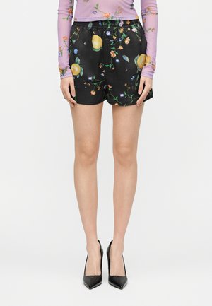 Femeie purtând pantaloni scurți negri cu model floral de lămâi, un top mov cu mâneci lungi și flori, și pantofi negri cu toc ascuțit, stând pe un fundal alb.