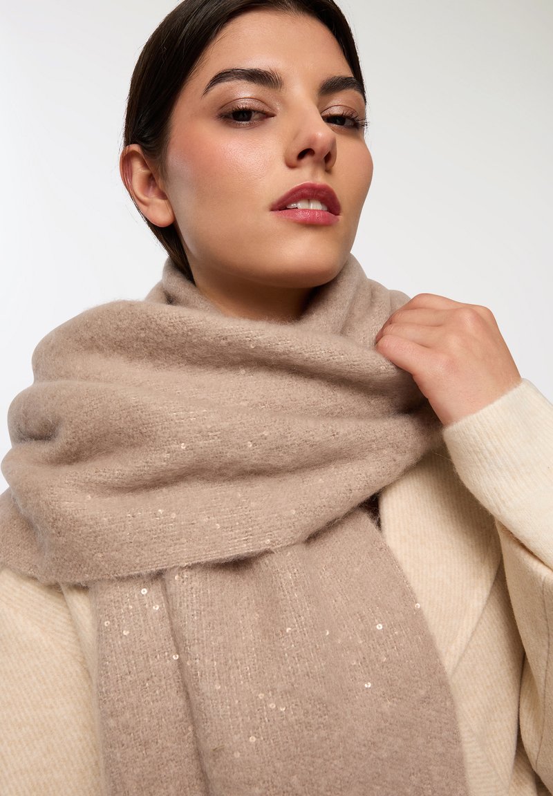 Sciarpa beige realizzata in un tessuto morbido, con delicate decorazioni, avvolta attorno al collo. Indossata con un maglione color crema.