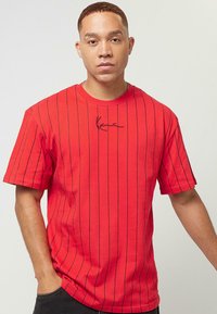 T-shirt en coton rouge avec des rayures verticales noires, manches courtes, col rond et un petit logo noir en signature sur la poitrine.