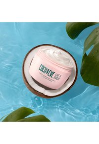 Roze pot met Coco & Eve haarmasker gehouden in een gehalveerde kokosnoot die drijft op helderblauw water, met groene bladeren in de buurt.