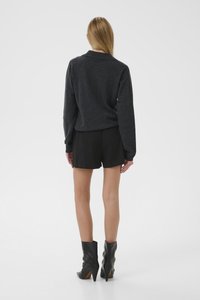 Mujer con cabello largo vistiendo un suéter gris oscuro, pantalones cortos negros y botas de tobillo con tacón negras, de pie contra un fondo blanco.