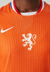 Orange atletisk tröja med texturerad tyg, med vertikala ränder och en vit lejonlogotyp på framsidan. Nike-logotyp i vitt på axeln.