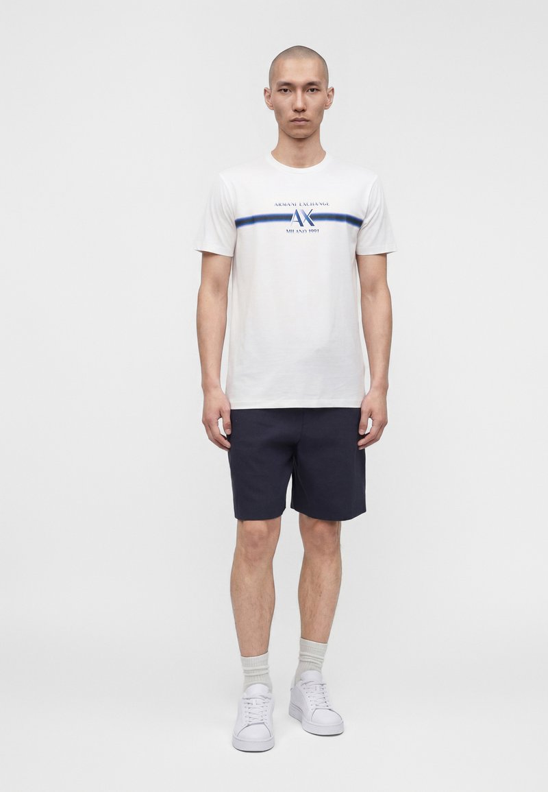 Homme debout portant un t-shirt blanc Armani Exchange avec un logo bleu, un short bleu marine, des baskets blanches et des chaussettes grises sur un fond clair uni.