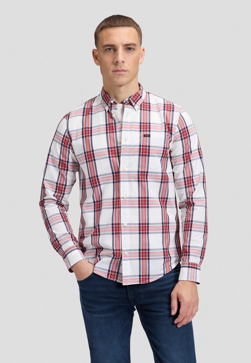 Homme portant une chemise à carreaux blanche, rouge et bleu marine à boutons, avec un jean bleu foncé, debout avec une main dans la poche sur un fond gris.