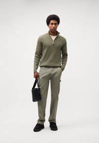 Oliven grøn strikpullover med halv lynlås, beige skjorte under, lys grøn bukser, sort taske og sorte sko med strukturerede såler.