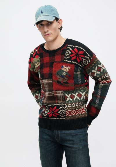 Joven vestido con un suéter de punto de patrón multicolor con un diseño de esquiador, jeans oscuros y una gorra de béisbol azul clara, de pie con las manos detrás de la espalda.
