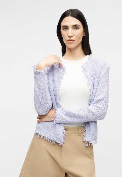 Νεαρή γυναίκα με μακριά σκούρα μαλλιά, φοράει ένα ανοιχτό μοβ υφασμάτινο ζακέτα πάνω από λευκή μπλούζα και μπεζ παντελόνι, στέκεται με το ένα χέρι κοντά στον ώμο της.