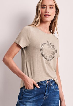 Donna che indossa una maglietta beige a maniche corte con motivo a puntini circolari e scritta "SOLEIL", abbinata a jeans blu, con la mano in tasca.