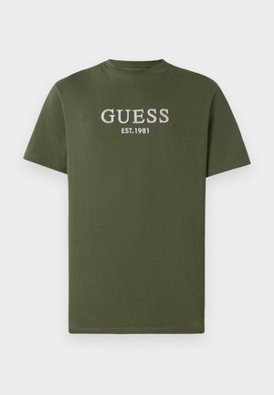 T-shirt en coton olive avec des manches courtes, arborant "GUESS EST. 1981" brodé en gris clair sur la poitrine. Design simple, col rond.