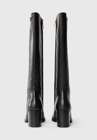 Bottes en cuir noir montant jusqu'au genou avec une texture lisse, fermeture éclair et talons blocs. Équipées de coutures verticales pour un design épuré.