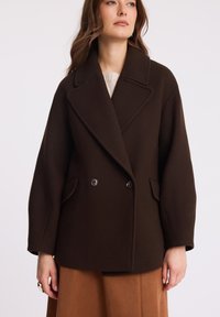 Podwójnie zapięty brązowy płaszcz wełniany z dużym zaokrąglonym kołnierzem, przednimi kieszeniami i guzikami jako akcentami. Gładka faktura i luźny krój.