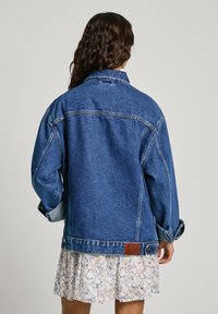 Denimjacka i medelblått med avslappnad passform, stor krage och knappade ärmar. Har läderpatchdetalj vid fållen.