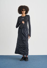 Mujer con cabello rizado que lleva una blusa negra de manga larga, falda larga negra, zapatillas negras, gafas de sol y un bolso cruzado, sobre un suelo azul.