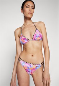 Bikini komplektas su gėlių raštu, kuriame dominuoja rožinė, violetinė ir mėlyna spalvos. Viršus turi trikampio formą; apatinė dalis turi dvigubas juostas su leopardiniu akcentu.