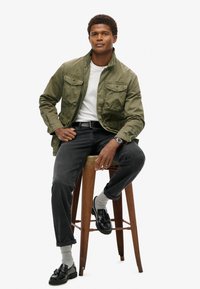 Veste légère olive avec deux poches poitrine, t-shirt blanc, jean noir et mocassins noirs, assis sur un tabouret en bois.