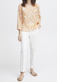 Una donna che indossa una blusa lunga a maniche lunghe di colore pesca, arancio e bianco, pantaloni bianchi su misura e sandali flat beige con punta aperta.