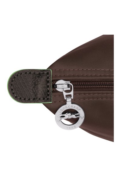 Brown fabric bag corner with a dark brown textured zipper pull, featuring a silver round charm embossed with "Longchamp 1948." Green accent.