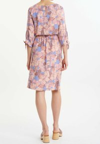 Kobieta stojąca tyłem, ubrana w sukienkę do kolan w kwiatowy wzór w różowe i fioletowe barwy oraz beżowe sandały na obcasie.