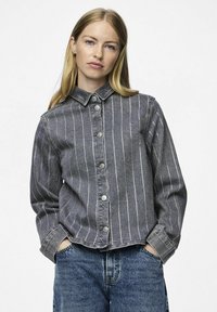 Giacca di jeans grigia con righe verticali bianche, chiusura con bottoni, colletto a punta e maniche lunghe. Indossata con jeans di denim blu.