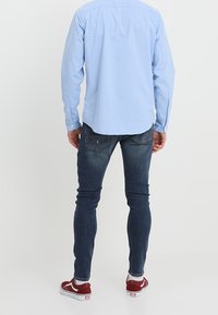 Muž oblečený v světle modré košili s dlouhým rukávem, tmavě modrých úzkých džínách, bílých ponožkách a červených teniskách stojí zády k bílé pozadí.