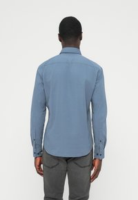 Μπλούζα μακριά μανίκια, ανοιχτό μπλε, με γιακά που κουμπώνει με κουμπιά, ευθεία βάση και μανσέτες με κουμπιά, κατασκευασμένη από ομαλό ύφασμα. Εμφανίζεται η πίσω όψη.