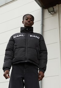 Giubbotto a vento nero con colletto alto, con testo del logo bianco "KARL KANI" sul petto; indossato con jeans scuri; texture liscia, design trapuntato.