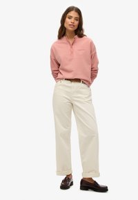 Camisa henley de manga larga color rosa claro con cuello y logo bordado, combinada con pantalones de puño enrollado color blanco roto y mocasines marrón oscuro.