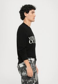Črn pleteni pulover z belim napisom "VERSACE COUTURE" spredaj; v kombinaciji z vzorčnimi črnobele hlače.