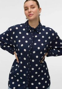 Chemise à manches longues bleu marine avec de grands pois blancs, fermeture à boutons sur le devant et col classique. Tissu texturé doux et poches latérales.